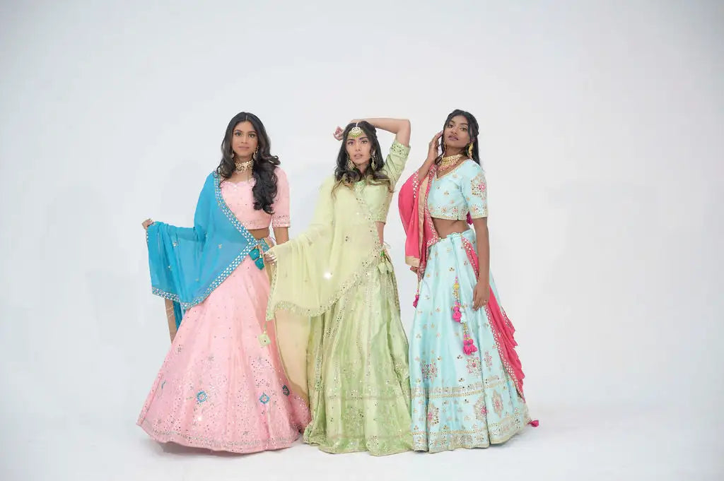 Three women are dressed in traditional indian lehengas, each featuring intricate embroidery and embellishments with distinct pastel colors, pink, green, and light blue, with matching dupattas or shawls.