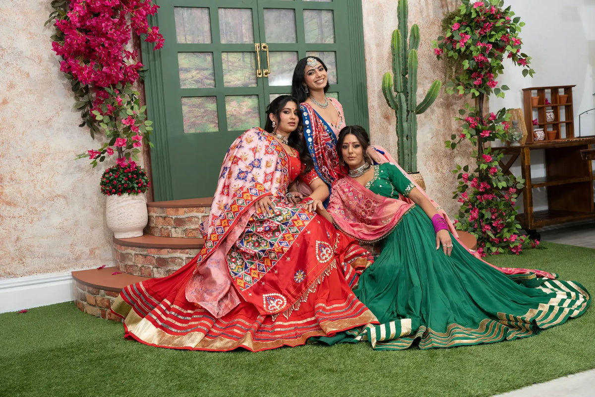 Three women are dressed in vibrant, intricately embroidered traditional indian lehengas with rich reds and greens.